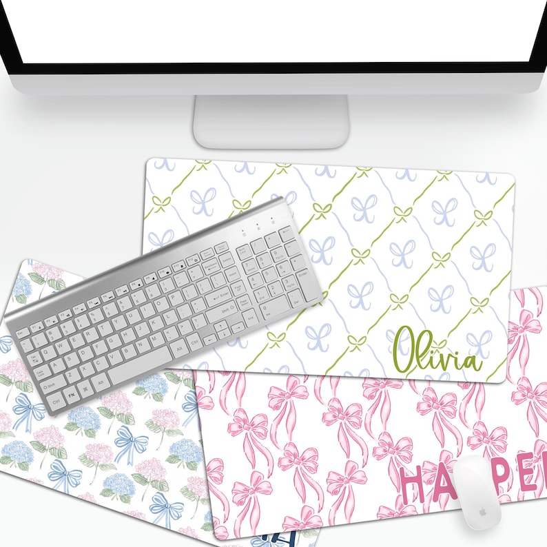 May include: Four different desk mats with various patterns. The mats feature a white background with different colored designs. The designs include blue and green bows, pink bows, and blue and pink hydrangeas. The mats are personalized with the names "Olivia" and "Harper".