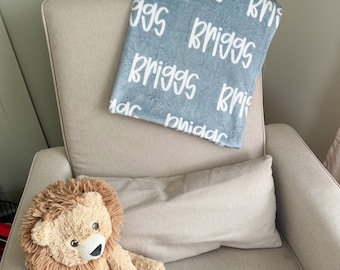 May include: A soft, light blue plush blanket featuring the name Briggs printed repeatedly in a white, handwritten-style font, accompanied by a tan, fuzzy stuffed lion toy with a brown mane, both resting on a neutral-colored upholstered chair.