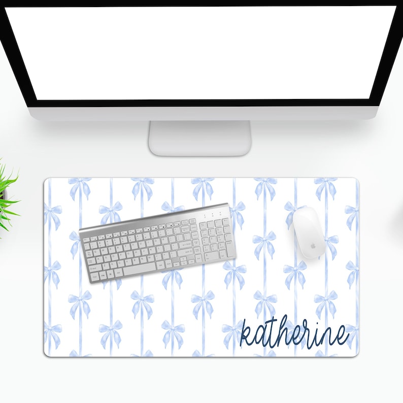 May include: A white desk mat with a light blue watercolor bow pattern. The mat has the name "katherine" written in dark blue cursive at the bottom. A silver keyboard and a white computer mouse are on the mat.