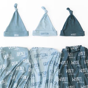 May include: Three baby hats in shades of blue, each with a knotted top and a folded brim. The hats are labeled with the names "Beau", "Grayson", and "Wyatt". Below are matching blankets with the names printed all over them.
