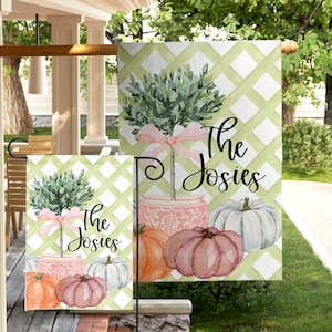 May include: A decorative garden flag featuring a watercolor design with a green trellis background. The flag includes a tree with a pink bow, pumpkins, and the text "The Josies".