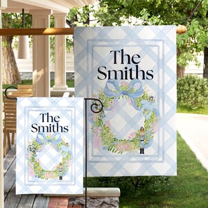 May include: Two garden flags with a light blue and white checked pattern. Each flag displays the text "The Smiths", a wreath with a blue bow, and floral details. The flags are displayed outside, likely in a garden.