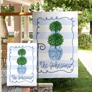 May include: A garden flag with a blue and white striped background. The flag features a green topiary in a blue and white patterned pot, with the text "The Johnsons" in a script font. A smaller matching flag is also visible.