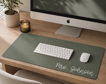 May include: A sage green desk mat with a white keyboard, mouse, and the text "Mrs. Johnson" in a cursive font. The mat sits on a wooden desk, with a computer monitor, a candle, and a small potted plant in the background.