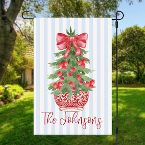 May include: A rectangular garden flag featuring a light blue and white vertical striped background. The center displays a watercolor Christmas tree in a red patterned bowl, topped with a red bow and red berries. The text The Johnsons is below.