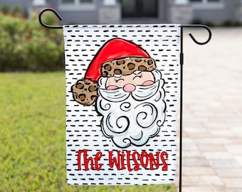 May include: A garden flag featuring a Santa Claus design. Santa wears a red hat with a leopard print brim and a white beard. The flag has a black and white dotted background and the name "The Wilsons" in red.