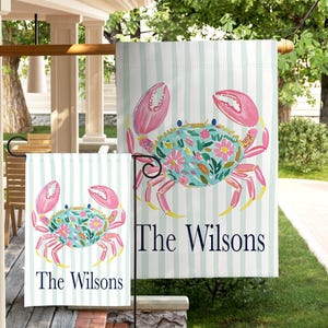 May include: A rectangular garden flag featuring a light blue and white vertical striped background with a central illustration of a crab decorated with pink flowers and green leaves, and the text The Wilsons printed in dark blue lettering.