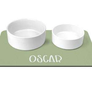 May include: A green pet food mat with the name "Oscar" printed on it. Two white ceramic bowls are on the mat.