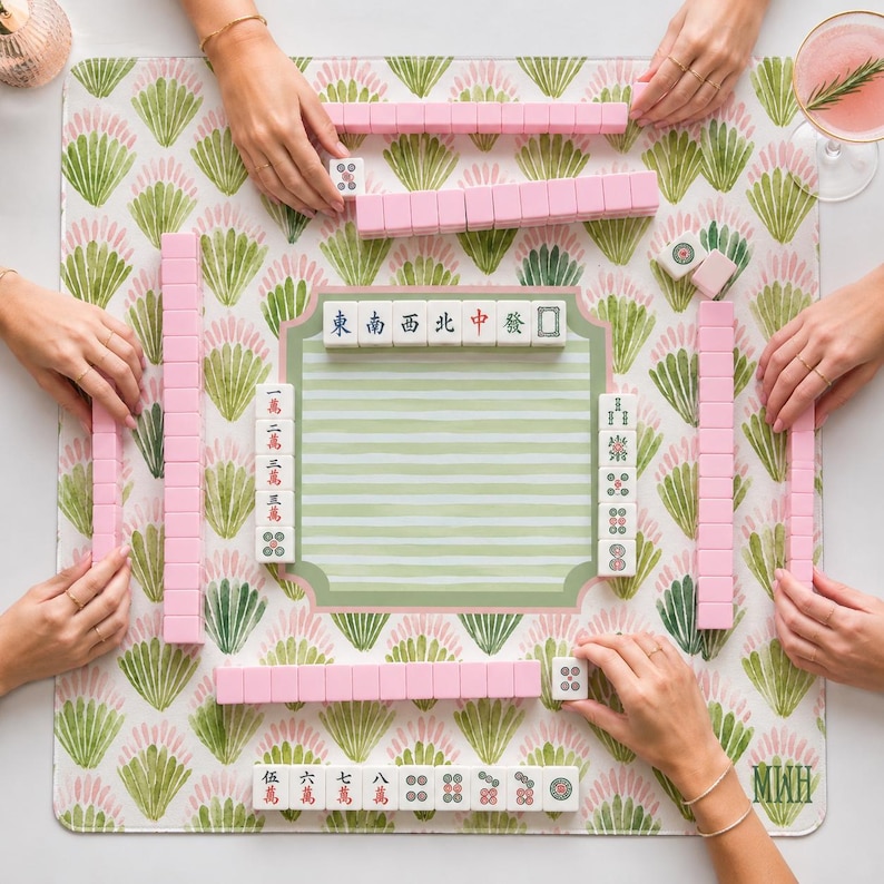 May include: A Mahjong game set featuring pink tiles with white faces, arranged on a square play mat decorated with a green and pink fan-shaped botanical pattern. The center of the mat displays a striped green area with various Mahjong tiles.