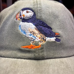 May include: A green baseball cap with an embroidered image of a puffin. The puffin is facing left and has a black, white, and orange beak. The puffin is white with black eyes and a black and blue back.