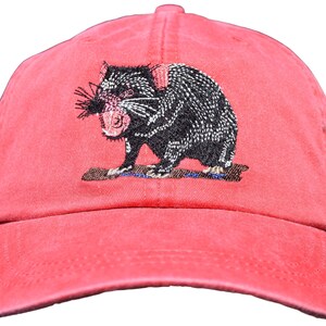 May include: A coral-colored baseball cap featuring an embroidered Tasmanian devil. The black and white devil has a pink mouth and is standing on a brown log. The cap has a curved brim and an adjustable strap.