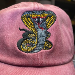 May include: A pink baseball cap with an embroidered cobra design. The cobra is green, yellow, and black.