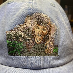 May include: A blue baseball cap with an embroidered image of a monkey's face. The monkey is brown and has a green background.