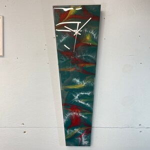 May include: A long, narrow, abstract clock with a white face and black hands. The clock face is set against a background of swirling green, red, and yellow paint.