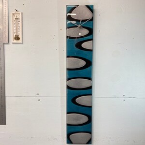 May include: A long, rectangular clock with a blue background and white and black abstract shapes. The clock face is in the center of the clock.