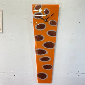 Puede incluir: Un reloj largo de vidrio naranja con manchas marrones. La esfera del reloj es blanca con manecillas negras y números negros.