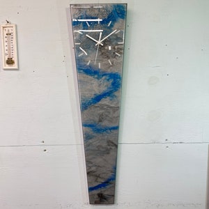 May include: A long, narrow, vertical clock with a white face and black hands. The clock is encased in a clear frame with a blue and gray abstract design.