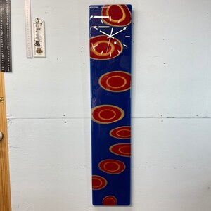 May include: A long, rectangular clock with a blue background and red and gold circles. The clock face is white with black hands.