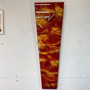 Peut inclure: Une horloge longue et étroite avec un design abstrait rouge, orange et jaune. Le cadran de l'horloge est blanc avec des aiguilles noires.