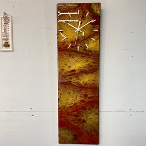 Peut inclure: Une horloge rectangulaire longue avec un cadran blanc et des aiguilles noires. L'horloge est placée sur un fond d'art abstrait rouge, jaune et orange.