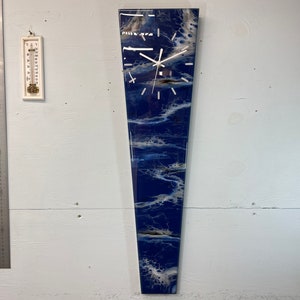 Puede incluir: Un reloj largo y estrecho, azul y blanco, con una esfera blanca y agujas negras. El reloj tiene forma de lágrima y un fondo blanco con remolinos azules y blancos.