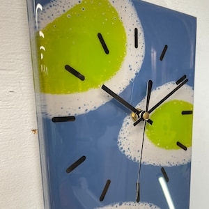 Puede incluir: Un reloj cuadrado con fondo azul y formas abstractas blancas y verdes. Las manecillas del reloj son negras y doradas.