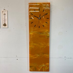 Könnte beinhalten: Eine lange, rechteckige Uhr mit einem gelben und weißen Marmorzifferblatt. Die Uhr hat schwarze Zeiger und schwarze Zahlen.