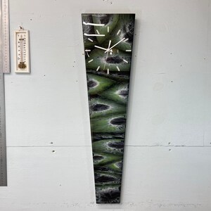 May include: A long, narrow, green and black clock with a white face and black hands. The clock is mounted on a white wall.