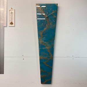 May include: A teal and gold resin clock with a white face and silver hands. The clock is shaped like a long, thin rectangle and is mounted on a white wall.
