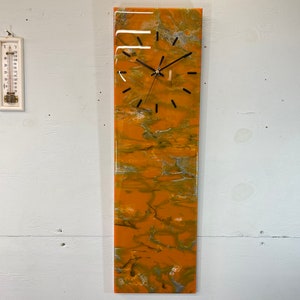 May include: A long, rectangular clock with a black face and black hands. The clock is mounted on a wall and has a background of orange, yellow, and white abstract art.