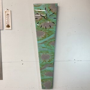 Peut inclure: Une horloge murale longue et abstraite avec un motif marbré turquoise et gris. L'horloge a des aiguilles et des index noirs. L'horloge mesure environ 122 cm de long.