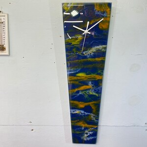 May include: A long, narrow, abstract clock with a white face and black hands. The clock face is set against a background of blue, yellow, and white resin.