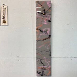 May include: A long, rectangular clock with a gray, pink, and black marbled design. The clock face is white with black hands.