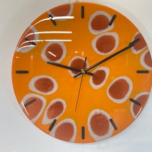 May include: A round orange clock with white and brown abstract designs. The clock has black hands and a gold centre.