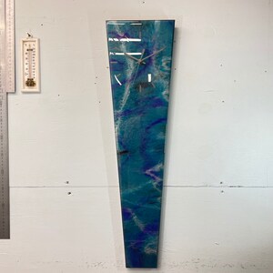 May include: A long, narrow, vertical clock with a white face and black hands. The clock is made of resin and has a blue, green, and purple abstract design.