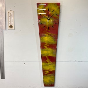 May include: A long, narrow clock with a red and yellow abstract design. The clock face is black with white hands and black numbers.