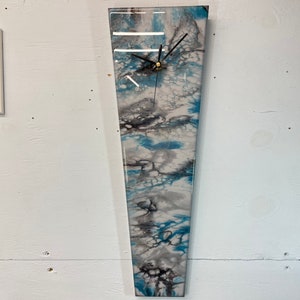 Puede incluir: Un reloj abstracto largo y estrecho, de color blanco, gris y azul, con agujas negras. La esfera del reloj es un vertido de resina con un diseño marmoleado blanco, gris y azul.