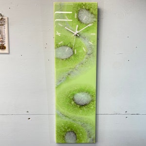 Peut inclure: Une horloge abstraite verte et blanche avec un cadran blanc et des aiguilles noires. L'horloge est montée sur un mur blanc.