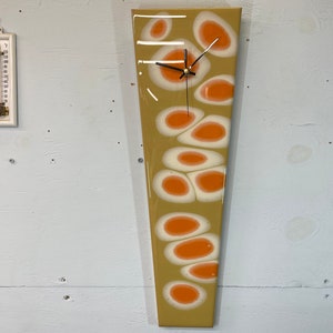 May include: A long, rectangular clock with a beige background and a pattern of orange and white circles. The clock has black hands and a gold centre.