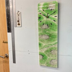 Può includere: Un orologio astratto verde e bianco con lancette nere. L'orologio è montato su una parete bianca.