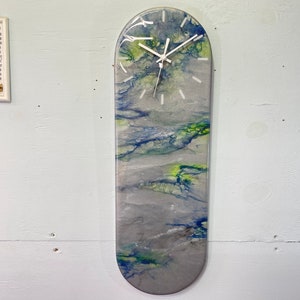 Może przedstawiać: Abstrakcyjny zegar w kolorze szarym, białym, niebieskim i zielonym z białą tarczą i czarnymi wskazówkami. Zegar ma kształt deski skateboardowej.