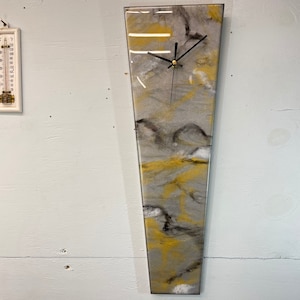 May include: A long, narrow, gray and yellow resin clock with black hands. The clock face is a light gray with a white background. The clock is hanging on a white wall.