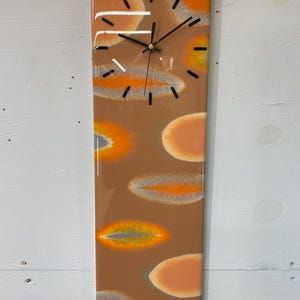 May include: A rectangular clock with a brown background and orange, yellow, and gray abstract shapes. The clock face has black hands and a gold center.