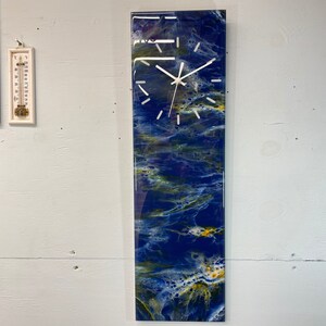 May include: A rectangular wall clock with a white face and black hands. The clock is set against a blue and yellow abstract background.