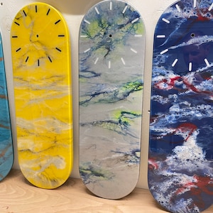 May include: Five skateboard-shaped clocks with different abstract designs. The clocks are painted in various colours, including blue, yellow, grey, and red. Each clock has a white face with black hands.
