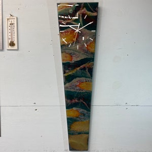 May include: A long, narrow, vertical clock with a white face and black hands. The clock is mounted on a wall and has a colorful abstract design with shades of green, blue, orange, and red.