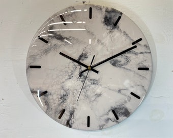 May include: A white and gray resin clock with black hands and black Roman numeral markings. The clock face has a marbled, abstract design.