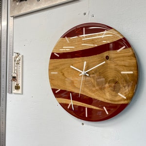 Może przedstawiać: Okrągły zegar drewniany z czerwonymi i białymi akcentami żywicznymi. Tarcza zegara ma białe wskazówki i białe oznaczenia godzin.