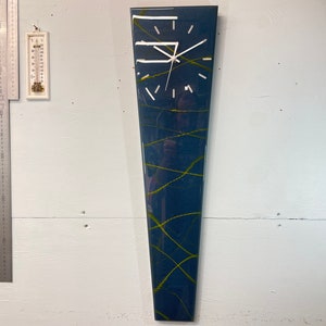 May include: A tall, narrow, blue and yellow clock with a white face and black hands. The clock is mounted on a white wall.