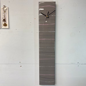 May include: A gray and white clock with a black face and hands. The clock has a unique design with thin pink lines running across the face. The clock is mounted on a wall.